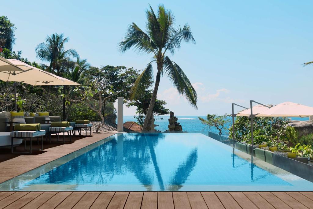 Eleganta bezgalības baseina ainava ar palmām un skatu uz jūru, radot relaksējošu atmosfēru. Terase ar modernas mēbeles papildina tropisko vidi.