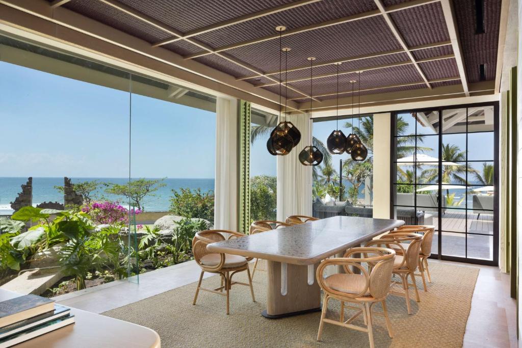 Mūsdienīgs ēdamistabas interjers ar skatu uz jūru, kurā ir liels galds un elegantas lampas. Apkārt ir zaļumi un terase ar palmām.