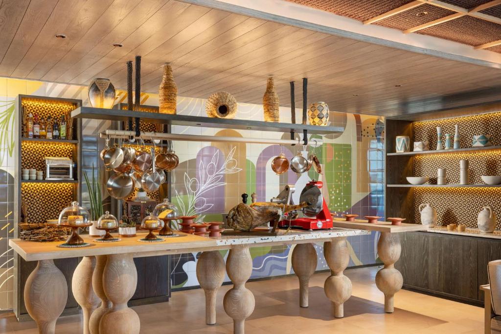 Moderns restorāns ar elegantu dizainu, kurā ir redzams liels galdiņš ar kulinārijas rīkiem, izsmalcināti trauki un dekoratīvi elementi uz krāsainas fona. Svaiga un mājīga atmosfēra.