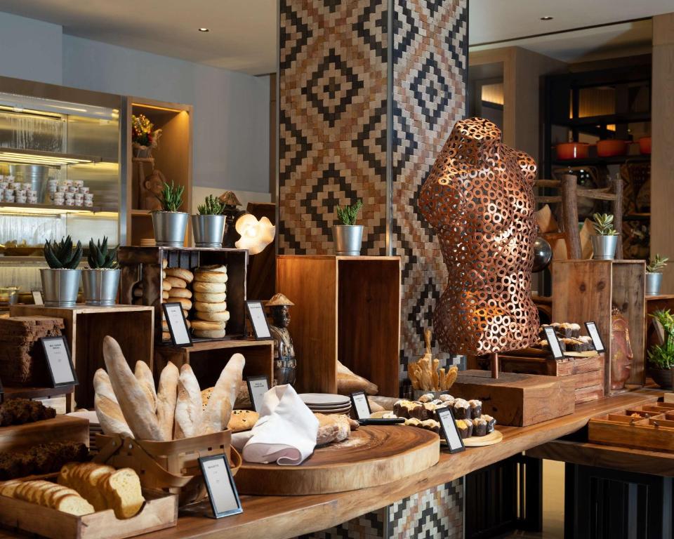 Kafejnīcas interjers ar dažādām maizēm un konditorejas izstrādājumiem, kas izkārtoti uz koka galdiem un plauktiem, papildināts ar dekoratīvām augu kompozīcijām un māksliniecisku skulptūru.