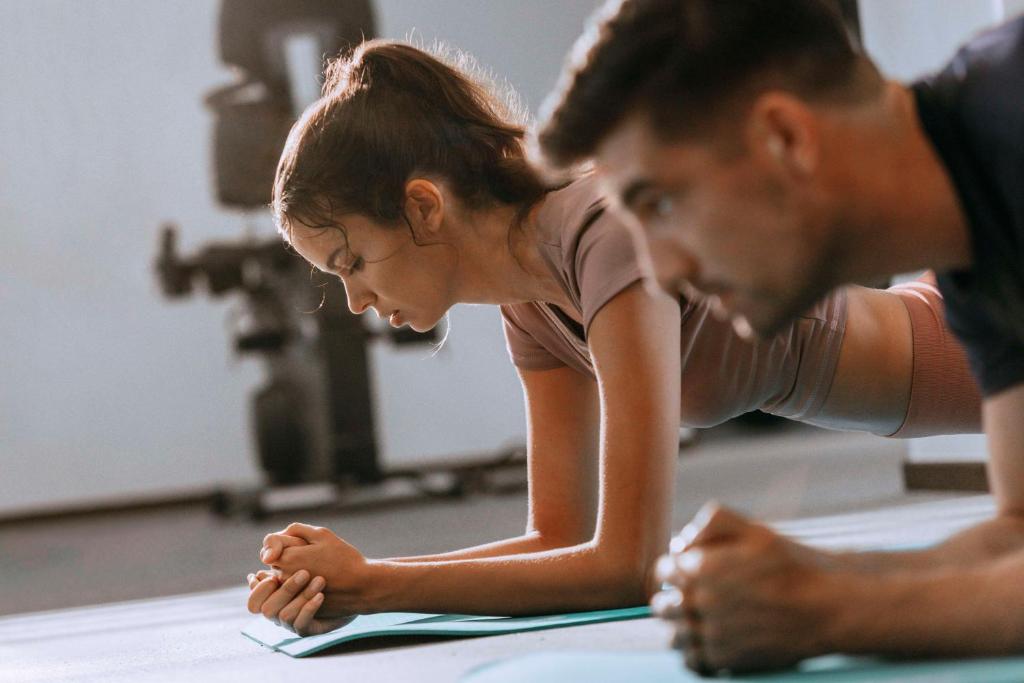 Treniņu sesija sporta zālē, kur divi cilvēki veic planku pozīciju uz vingrošanas paklāja. Ievērojama uzmanība un koncentrācija.