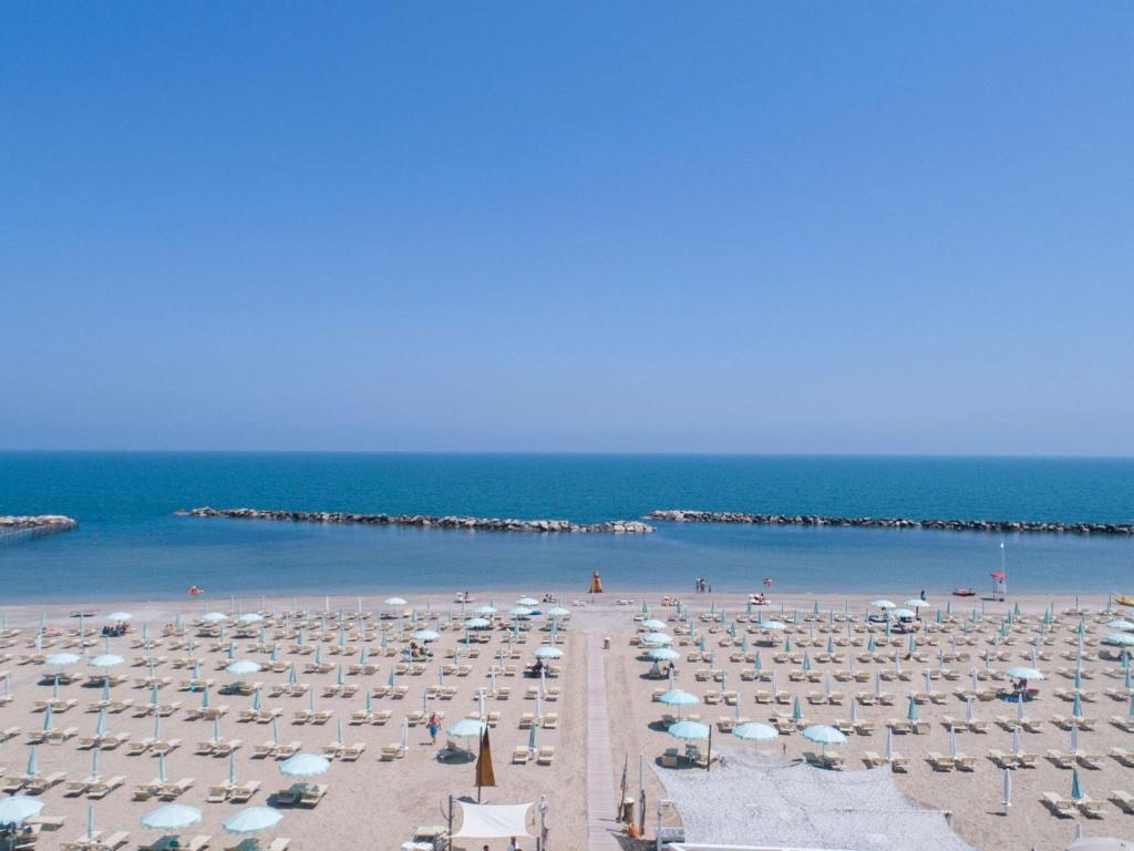 Pludmale ar balto smilšu un zilu jūru, kurā redzami sauļošanās krēslu un saulessargu rindu. Skats uz mierīgu ūdeni un skaidru, zilu debesi.