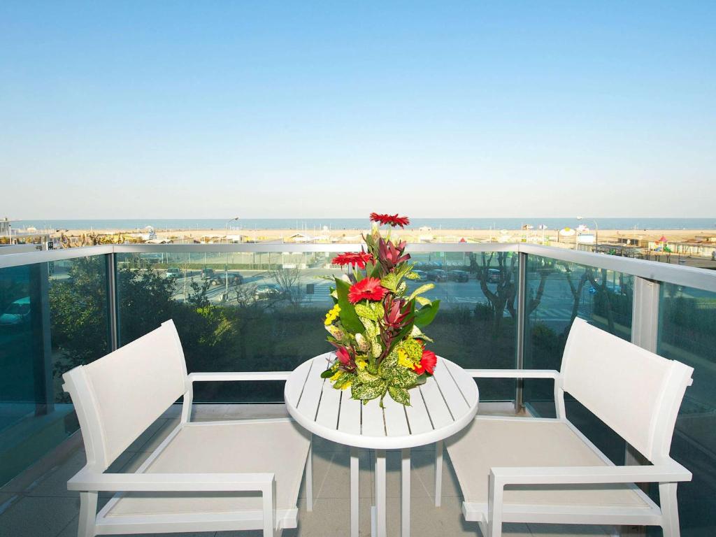 Balkona skats uz jūru ar ziedu kompozīciju uz galda. Balti krēsli un galds piedāvā mierīgu atpūtas vietu.