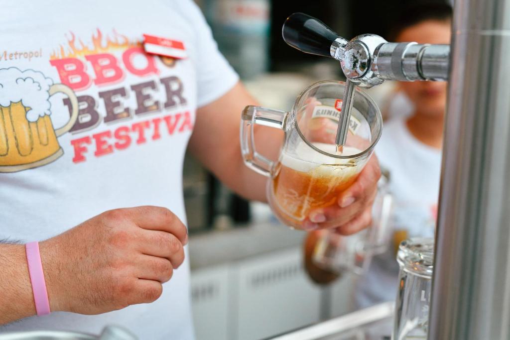 Vīna izsniegšana festivālā, kurā redzams bārmenis ar T-kreklu, uz kura ir uzraksts "BBQ Beer Festival", ielejot alu caur krānu.