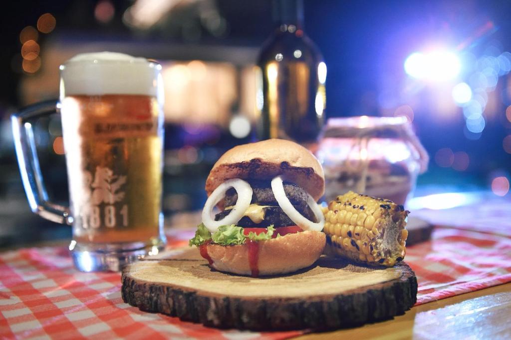 Svaigs burgeris ar dārzeņiem un alu, kas pasniegts uz koka šķīvja, ar grauzdētu kukurūzu un romantisku nakts apgaismojumu fonā.