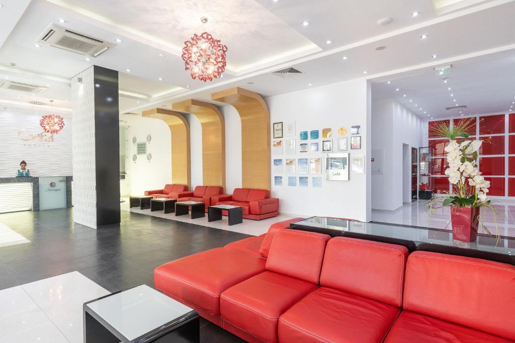 Mūsdienīgs viesnīcas recepcijas interjers ar sarkaniem dīvāniem, stilīgu apgaismojumu un dekoratīvām sienām. Redzams viesmīlis pie reģistrācijas letes.
