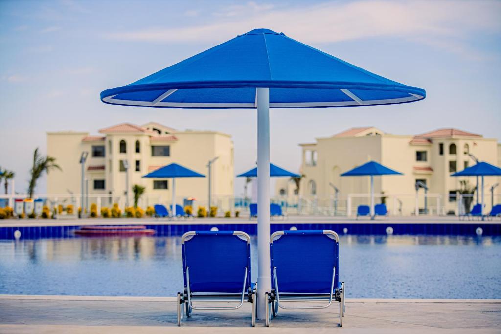 Divas zila krēsli ar lietussargu pie baseina, fonā redzami balti ēku kompleksi un zils ūdens.