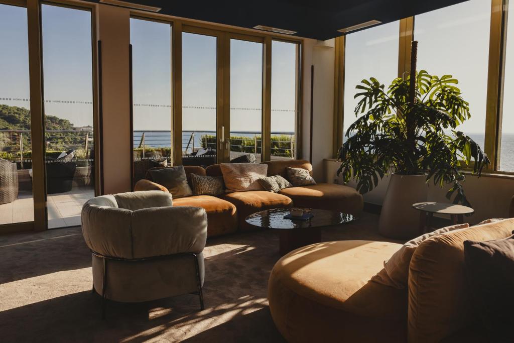 Mūsdienīgs viesistabas interjers ar plašiem logiem, caur kuriem redzama jūras ainava. Telpā ir ērti dīvāni, liels kafijas galdiņš un zaļa augšana, radot mierīgu atmosfēru.