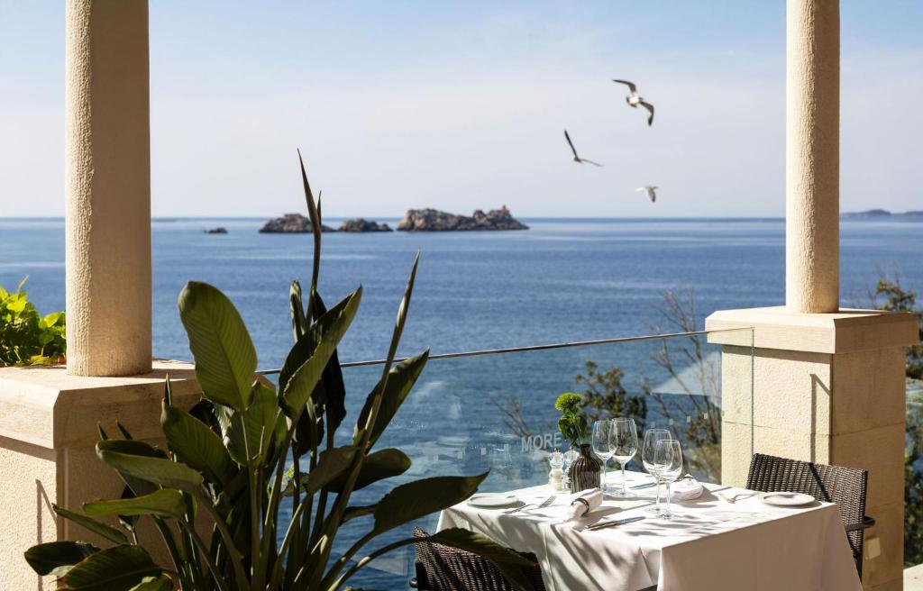 Skats uz jūru no restorāna terases ar elegantu galdu, kas klāts ar glāzēm un ziediem, un putni, kas lido pāri ūdenim.