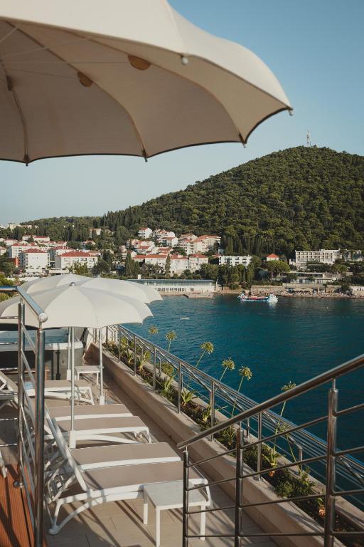 Skats no terases ar sauļošanās krēsliem un saulessargiem, kas vērsti uz jūru un kalnu ainavu. Fons papildina apburoša piekrastes pilsēta ar zaļām kalnu nogāzēm.