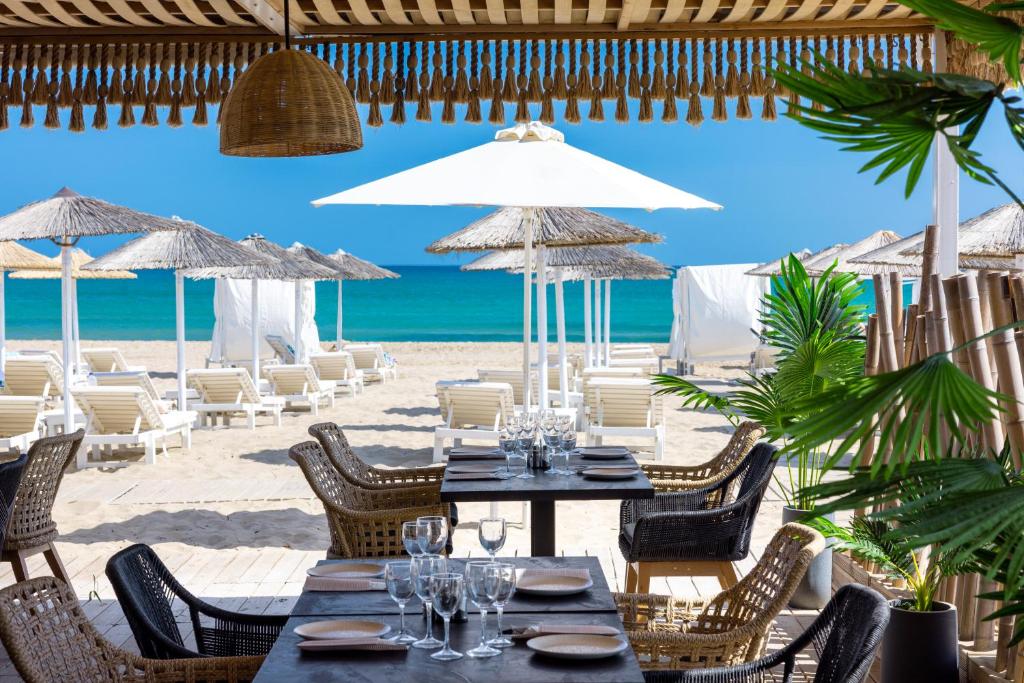 Pludmales restorāns ar skatu uz jūru, kur galdos sakārtotas glāzes un trauki, un fons, kurā redzamas sauļošanās krēsli un saulessargi.