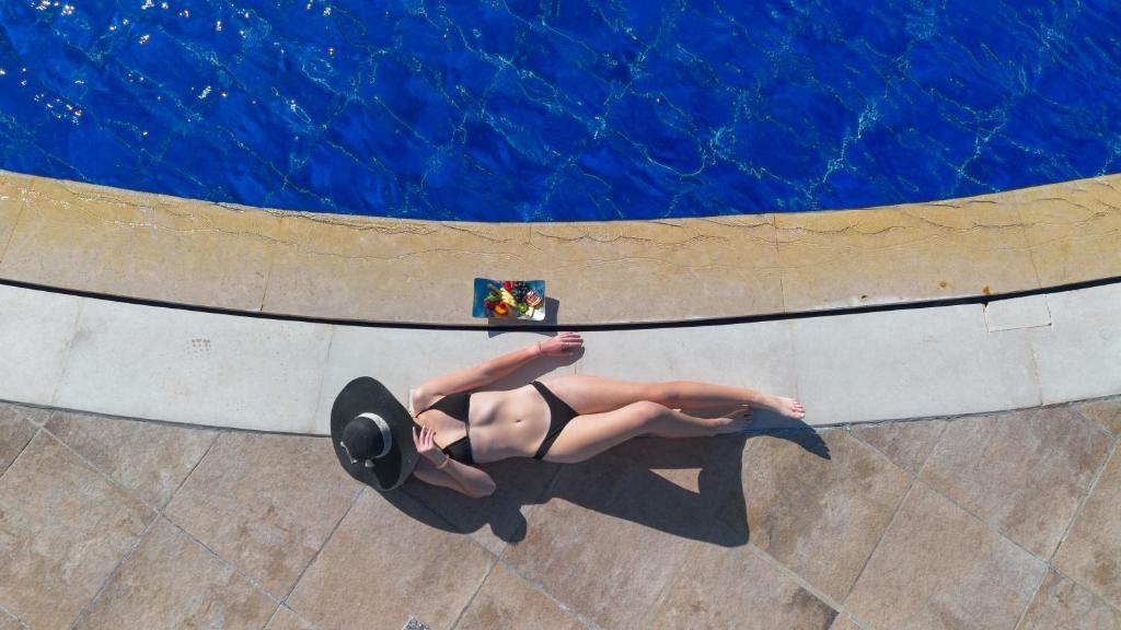 Saulesgaismošana pie baseina ar sievieti melnā peldkostīmā, kas atpūšas uz akmeņainas virsmas un blakus ir augļu šķīvis.