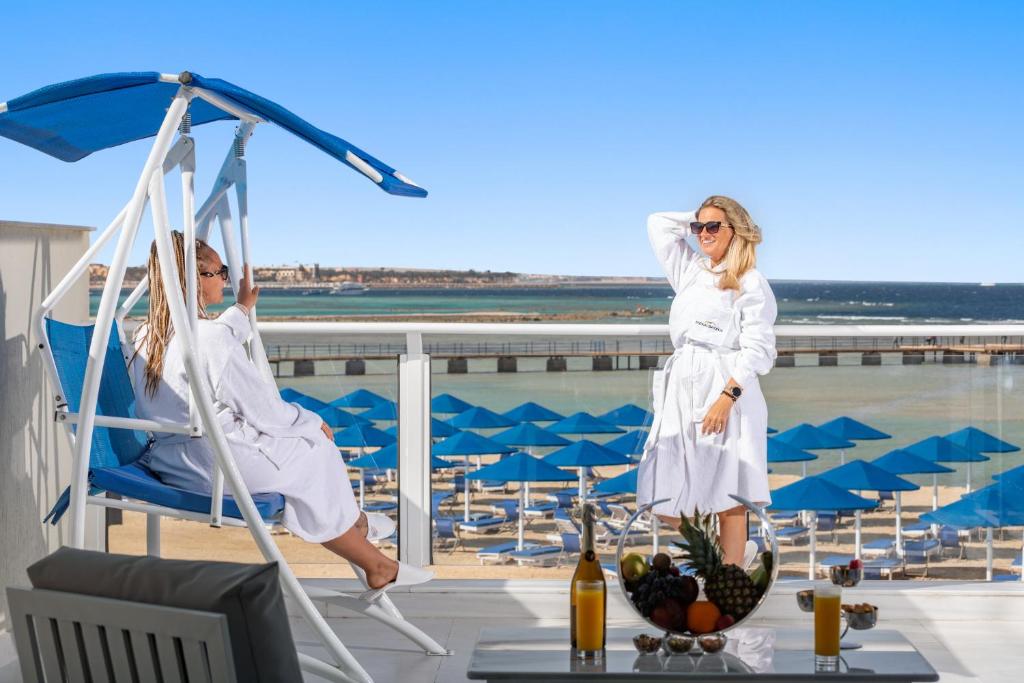 Divas sievietes baltos halātos baudīšanas mirklī uz terases ar jūru un pludmali fona, pie galda ar augļiem un dzērieniem. Ideāla brīvdienu atmosfēra.