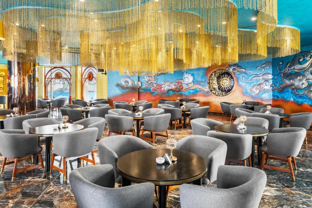 Moderns restorāns ar elegantu interjeru, kurā ir stilīgas pelēkas krēsli un melni galdi, apgaismoti ar mirdzošām zelta piekārtnēm un krāsainu sienu gleznojumu.