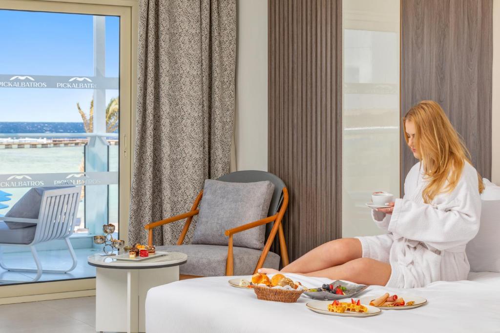 Eleganta viesnīcas istaba ar skatu uz jūru, kur jauna sieviete ģērbta halātā bauda kafiju un brokastis, kas pasniegtas uz gultas. Interjers ir gaišs un mūsdienīgs, piedāvājot relaksējošu atmosfēru.