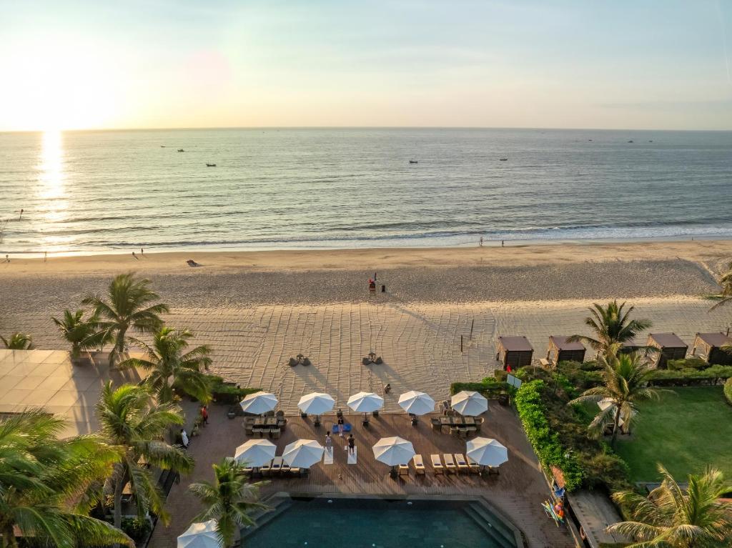 Pludmales ainava ar saulrietu, kur skatāma jūra un pludmale, apzaļumotas teritorijas ar sauļošanās krēsliem un lietussargiem.