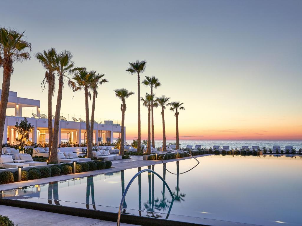 Grecotel Lux Me White Palace baseins ar palmām piekrastē saulrieta laikā.