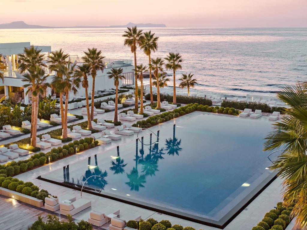 Grecotel Lux Me White Palace baseins piekrastē ar palmām un saulrietu.