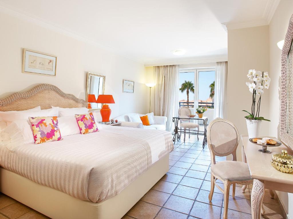 Grecotel Casa Adele numurs ar gultu un mēbelēm, saules gaisma iekļūst caur logiem.