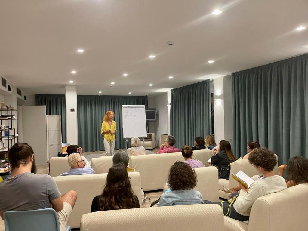 Seminārs, kurā dalībnieki klausās runātāju, kas stāv pie tāfeles. Telpa ir gaiša un moderna, ar zaļām aizkariem un ērtiem dīvāniem.