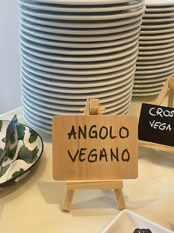 Koka zīme ar uzrakstu "ANGolo VEGANO" uz galda, fonā redzami sakrauti šķīvji un divi trauki ar ēdienu.