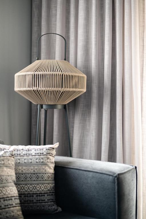 Mūsdienīgs lampas dizains ar ģeometrisku formu un pelēkām aizkariem, kas piešķir telpai elegantu izskatu. Sofas spilveni ar rakstainu tekstūru papildina interjera estētiku.