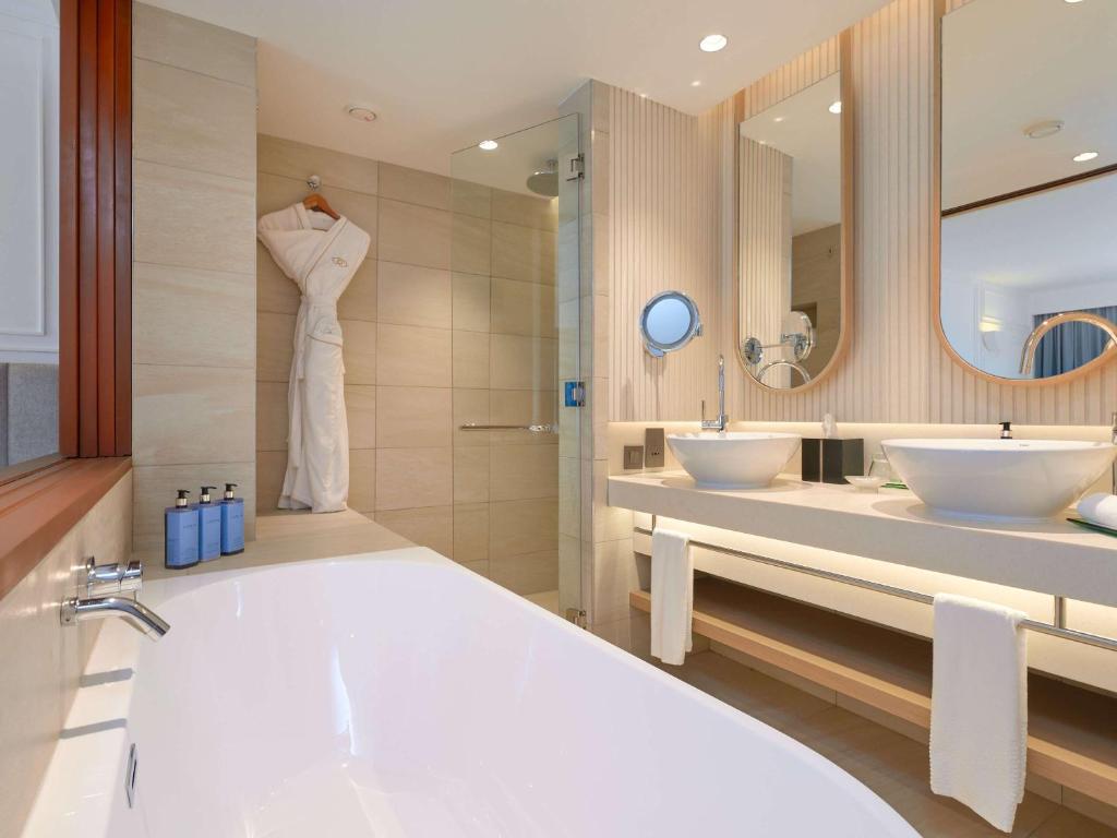 Moderns vannas istabas interjers ar elegantu vannu, divām izlietnēm un atspoguļojošām virsmām. Svaigi balta halāta un higiēnas līdzekļu komplekts rada komfortablu atmosfēru.