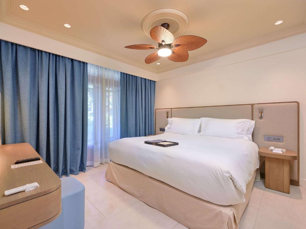 Mūsdienīgs guļamistabas interjers ar plašu gultu, zilu aizkaru un stilīgu ventilatoru. Labi apgaismota telpa ar koka mēbelēm un elegantu dizainu.