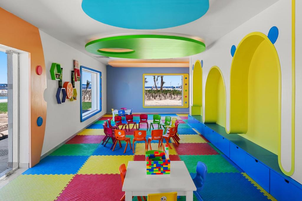 Krāsaina bērnu istaba ar daudzkrāsainu grīdu un zemu galdu, ap kuru ir sēdvietas. Logi piedāvā skatu uz āru, un telpā ir dažādas rotaļlietas un dekorācijas.
