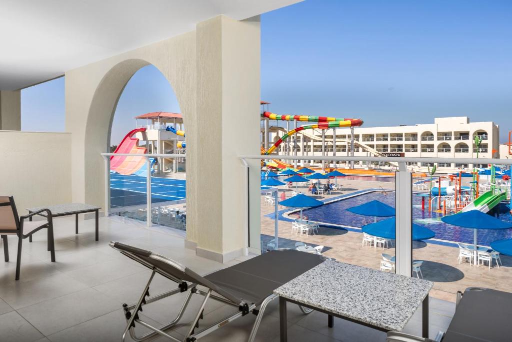 Terase ar skatu uz ūdens atrakciju parku, kur redzami ūdens slidkalniņi un baseins, ar sauļošanās krēsliem un ziliem saulessargiem.