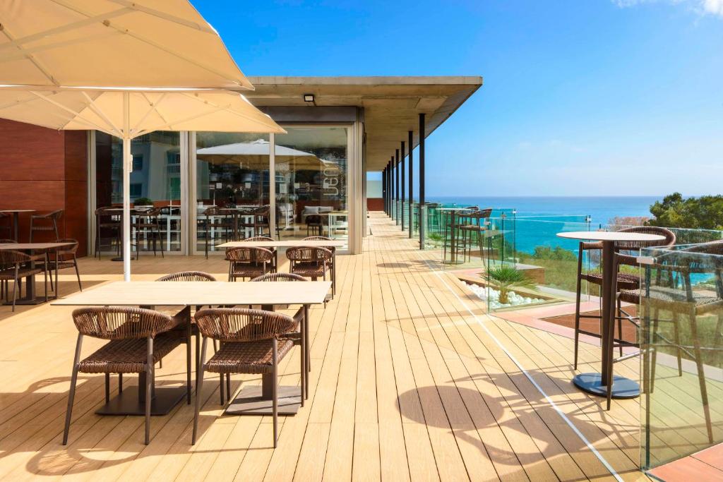Mūsdienīgs terases restorāns ar skatu uz jūru, kurā ir eleganti galdi un krēsli, kā arī saulessargi, kas piedāvā komfortu. Šī vide ir ideāla relaksācijai un baudīšanai brīvā dabā.