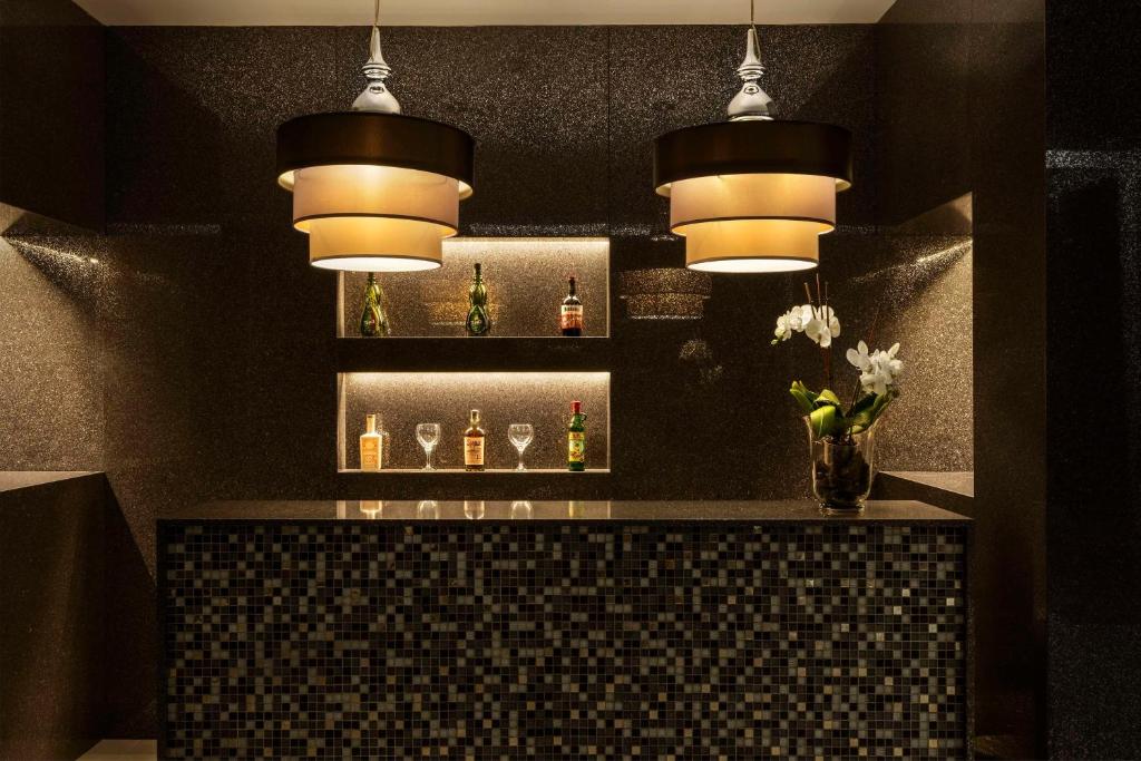 Mūsdienīgs bārs ar elegantiem lampām un tumšu fonu, kas izceļ dzērienus un glāzes uz plaukta. Apstādījumi pie bāra piešķir siltumu un stilu.