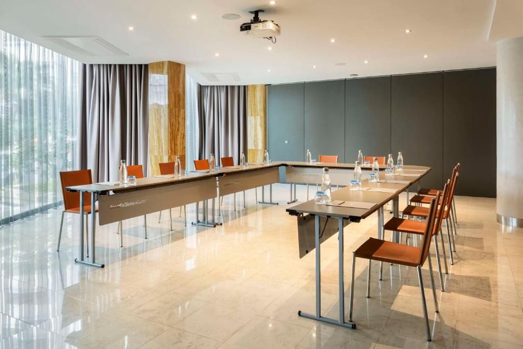 Mūsdienīga konferenču telpa ar garu galdu un krēsliem, kas aprīkota ar ūdens pudelēm un projektoru. Telpa ir apgaismota ar dabīgu gaismu no logiem un ir eleganta ar neitrāliem toņiem.