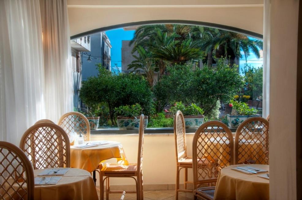 Gaisma piepildīta restorāna iekārtojums ar koka krēsliem un galdiem, kas vērsti uz skaistu dārzu ar palmām un ziediem. Logi atver skatu uz mierīgu ārpusi, radot viesmīlīgu atmosfēru.