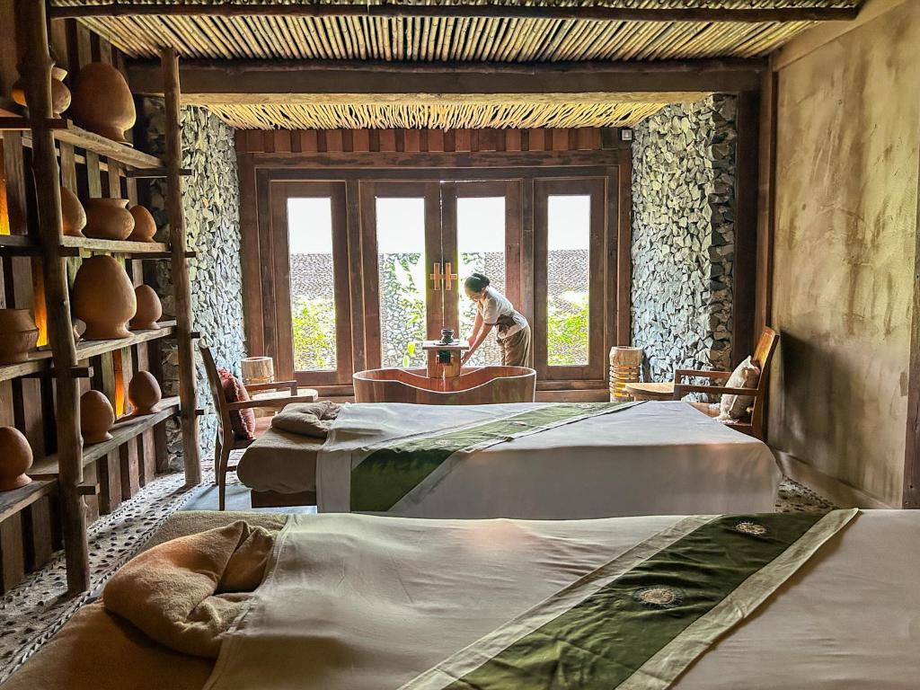 Spa telpa ar divām masāžas gultām, koka un akmens elementiem, un darbinieku, kas sagatavo telpu. Dabas gaisma plūst caur lielajiem logiem.