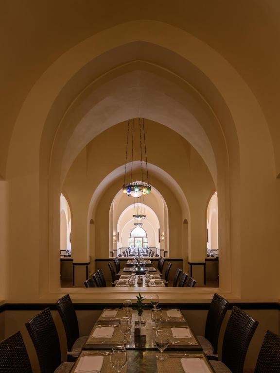 Eleganta restorāna interjers ar izsmalcinātiem arku veida griestiem un garām galdu rindām, kas rotātas ar glāzēm un ēdienu piederumiem. Atmosfēra ir mierīga un gaumīga, radot viesmīlīgu vidi.