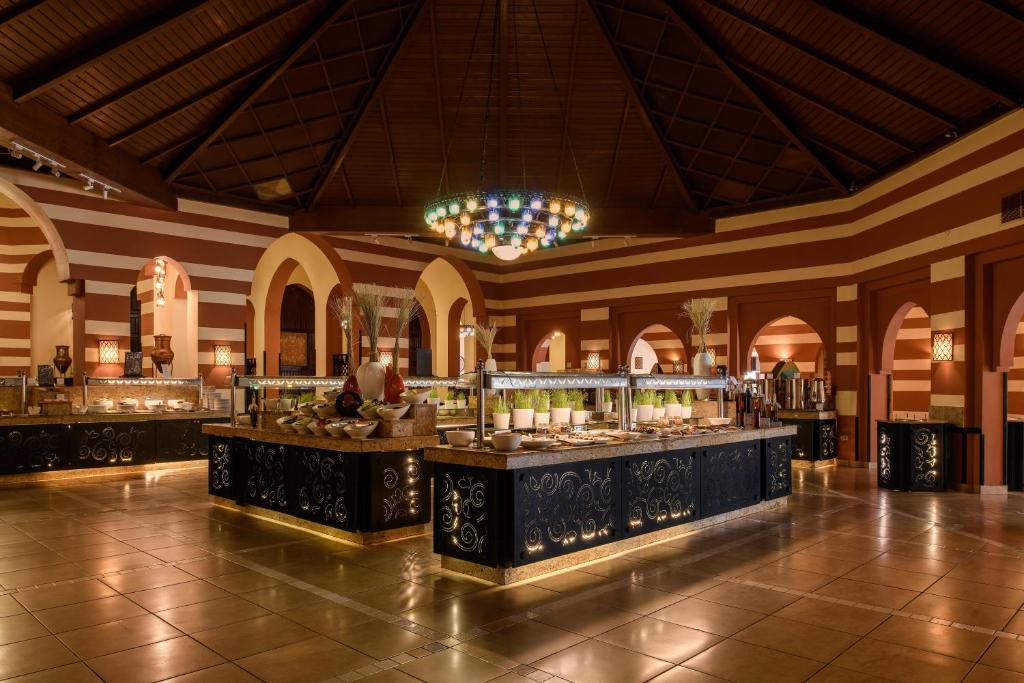 Elegantā restorāna interjers ar plašu bufetes zonu, kas rotāta ar krāsainu lampu un izsmalcinātu dizainu. Apgaismojums un arhitektūra veido siltu un viesmīlīgu atmosfēru.