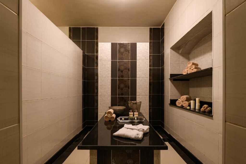 Mūsdienīga spa telpa ar melnu akmens galdu, baltām flīzēm un uzglabāšanas plauktiem, kuros ir salocītas dvieļi un sveces.