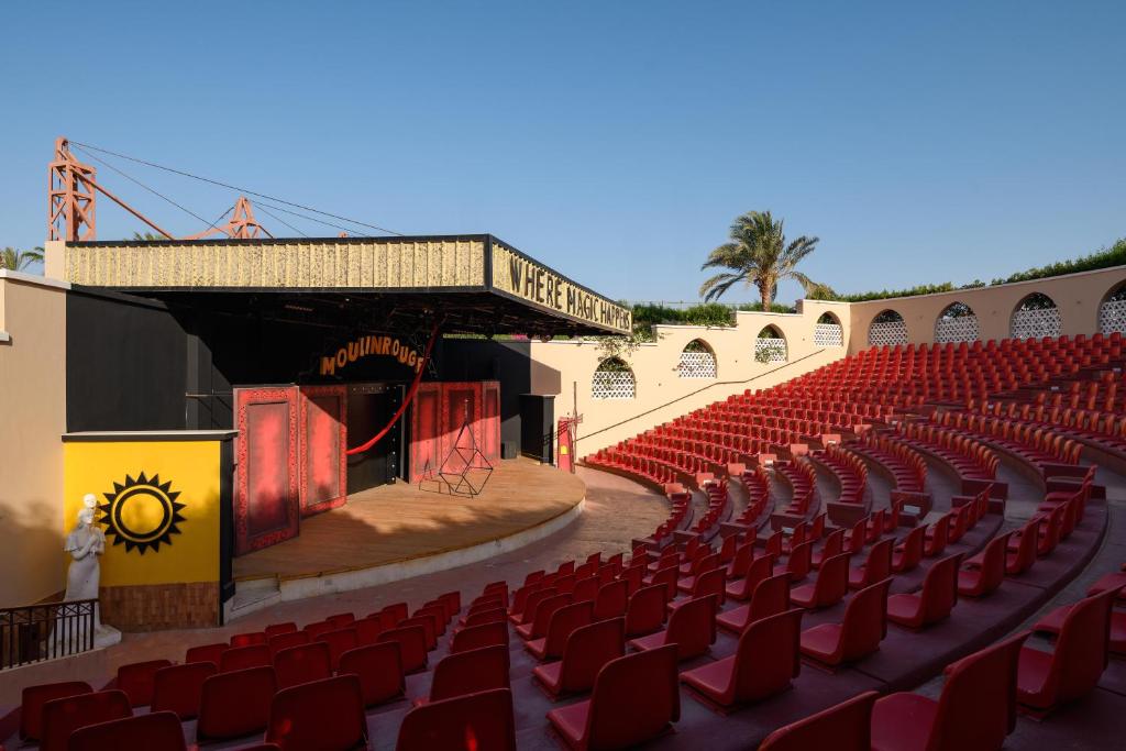 Āra teātris ar sarkanām krēsliem un skatuvi, kurā redzama zīme "Moulin Rouge" un "Where Magic Happens". Apkārt palmām un skaidrām debesīm.