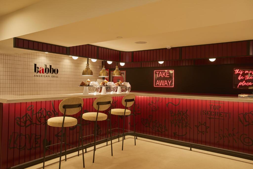 Mūsdienīga restorāna interjers ar atvērto bāru, sarkanu apdari, gaismas piekari un neonizētiem uzrakstiem "TAKE AWAY". Bāra krēsli un dekoratīvas detaļas veido viesmīlīgu atmosfēru.