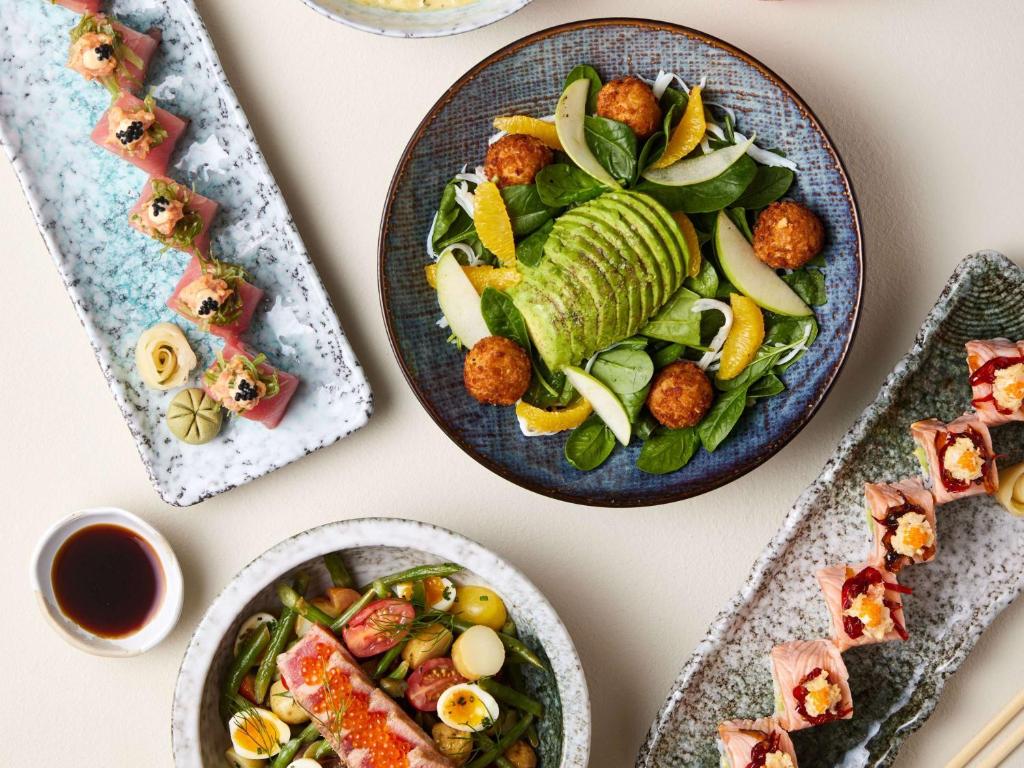 Svaigu sushi un garšīgas uzkodas, ieskaitot avokado salātus, rīsu bumbiņas un dārzeņus, pasniegti elegantās šķīvju kolekcijās.