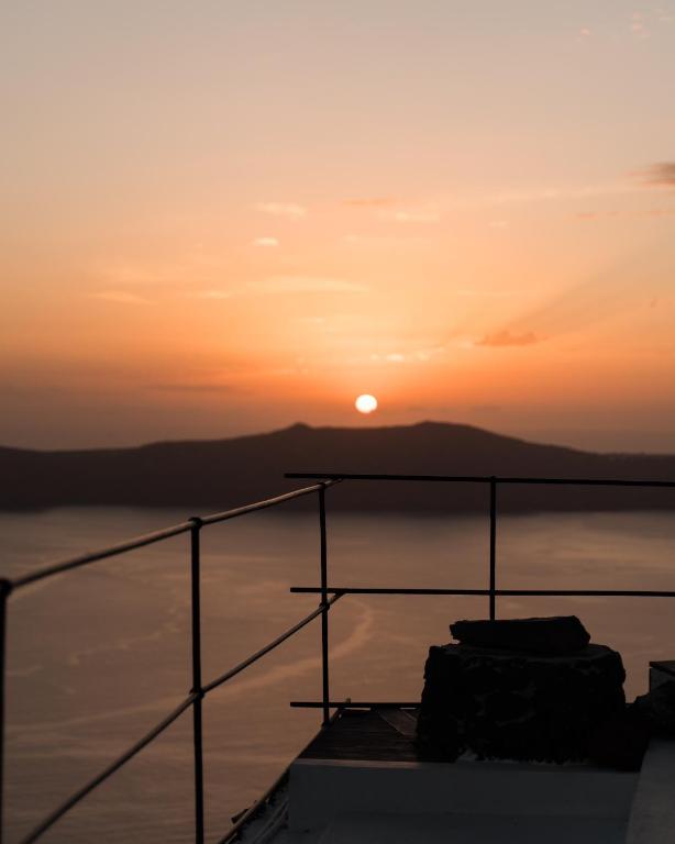 Saulriets virs kalniem ar skatu uz jūru, apvīts siltā oranžā gaismā. Rāmis ar metāla margām piešķir attēlam dziļumu.