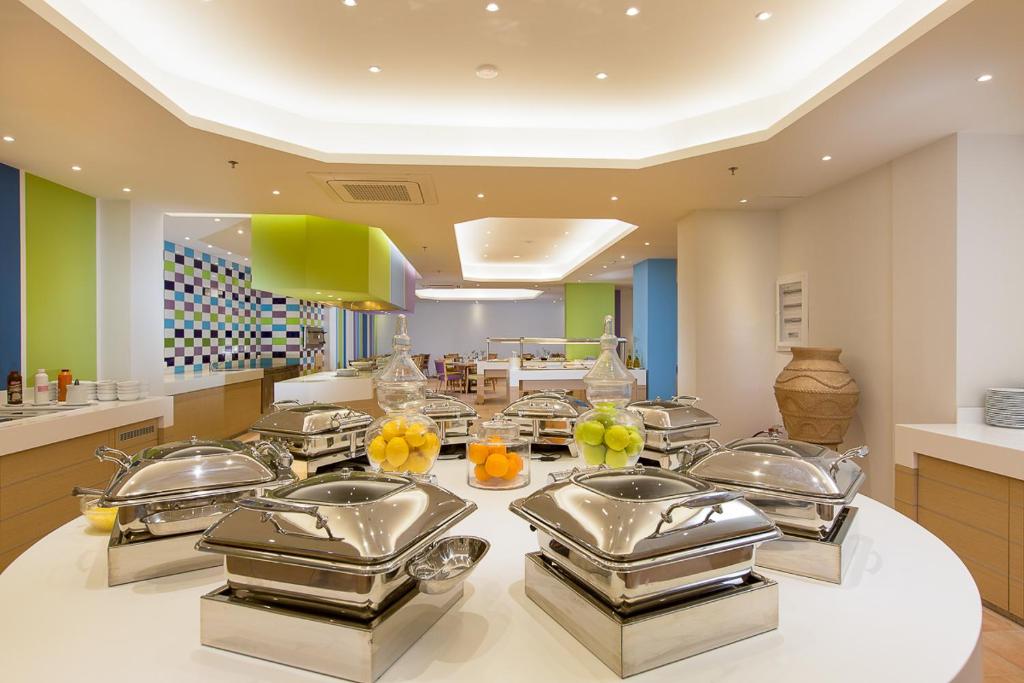 Gaiša un moderna bufete ar spīdīgiem traukiem un krāsainiem sienu elementiem, kurā redzami augļi un dažādi ēdieni. Ideāla vide brokastīm vai pusdienām.