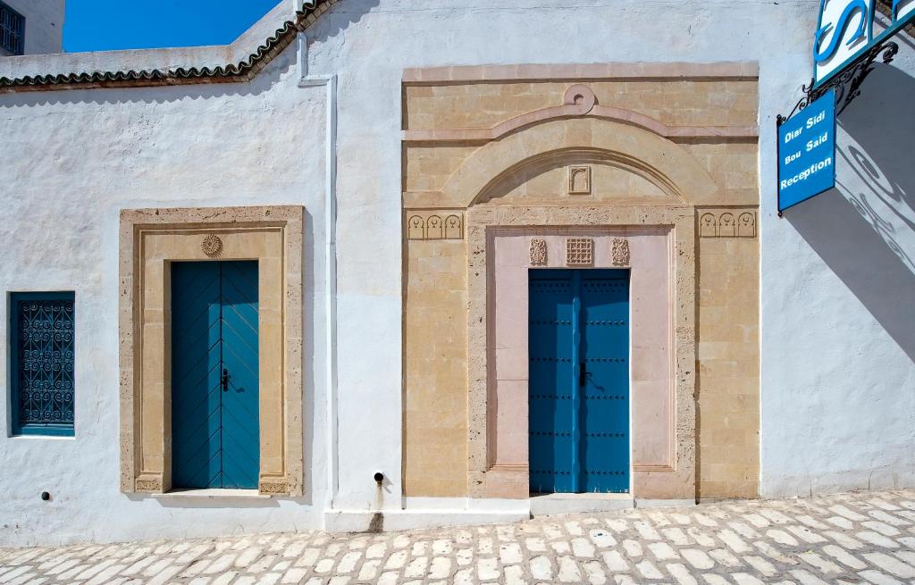 Tradicionālas arhitektūras durvis ar zilu krāsu un akmens apdari, kas atrodas tunisiešu pilsētā. Attēlā redzamas divas durvis, kas papildina vietas kultūras noskaņu.