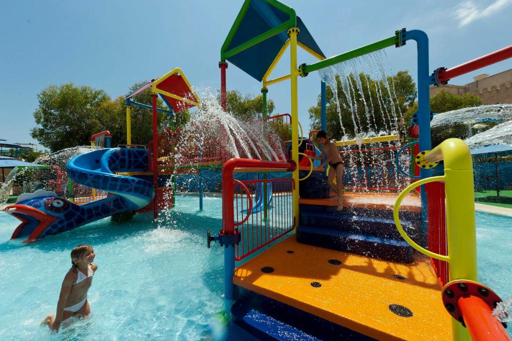 Bērnu ūdens atrakciju parks ar slidkalniņu un ūdens strūklakām, kur bērni spēlējas un izbauda ūdens prieku. Brightly colored structures and playful elements create a fun atmosphere.