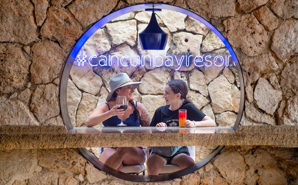 Divas sievietes, kas izbauda dzērienus Cancun Bay Resort bārā ar akmens sienu fonā.