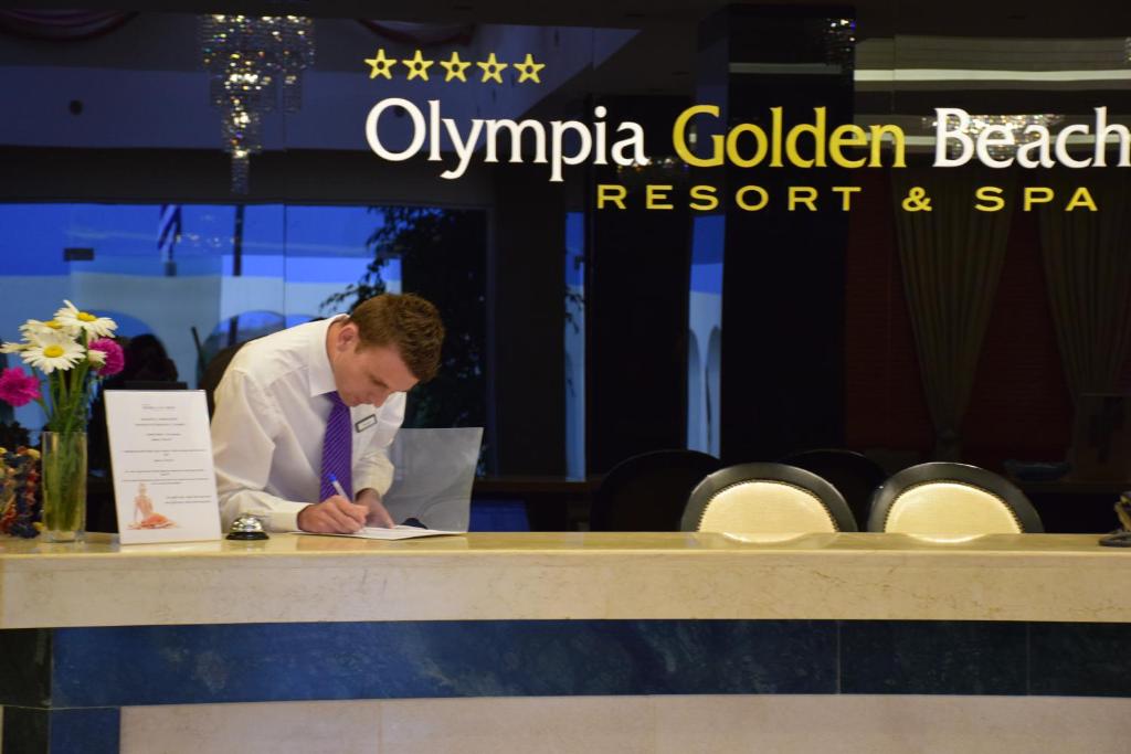 Reģistratūras darbinieks pieņem viesus Olympia Golden Beach Resort & Spa. Uz galda ir ziedi un informatīvi materiāli.