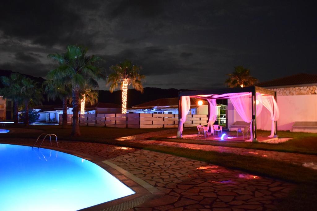 Nakts ainava ar baseinu un palmām, apgaismota ar krāsainām gaismām. Galds un krēsli atrodas zem nojumes, radot relaksējošu atmosfēru.