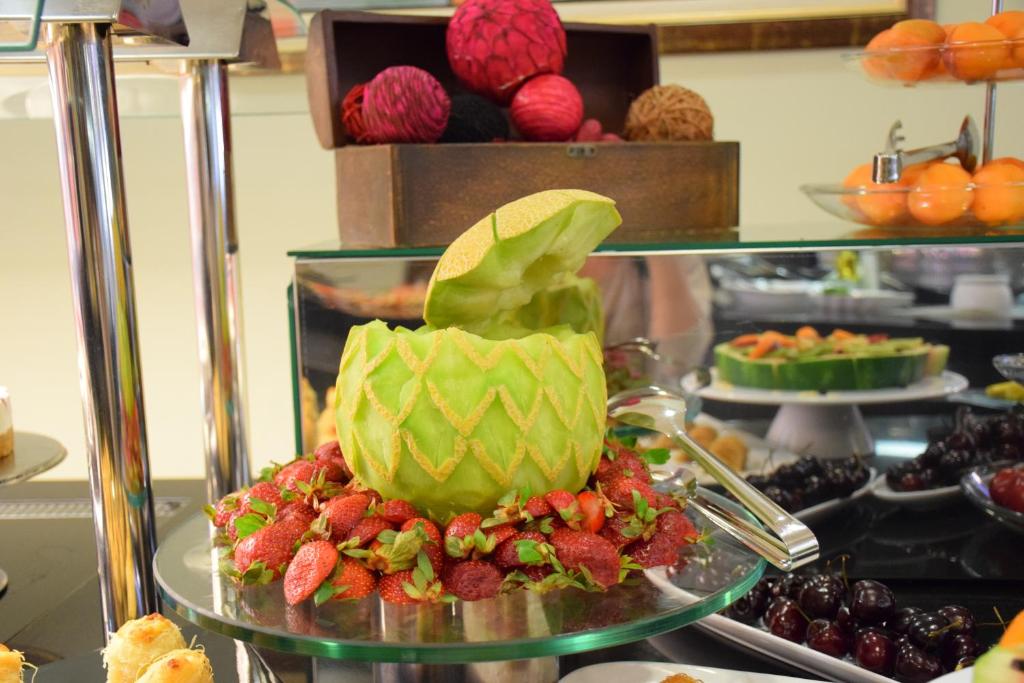 Krāšņs augļu galds ar dekoratīvu melone, kas izgriezta kā ananāss, apņemta ar zemenēm un citām ogām. Ideāls piemērs svinību dekorēšanai un viesību galda estētikai.