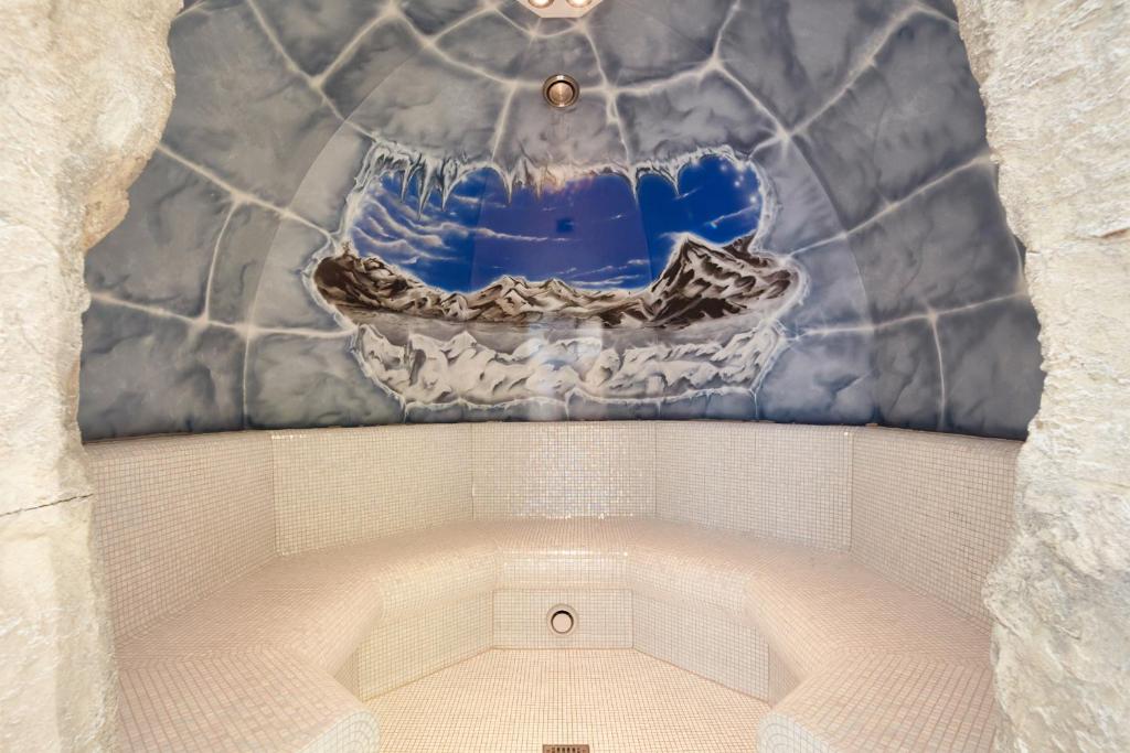 Siltā tvaika pirts ar kalnu ainavu uz sienas, veidota ar plūstošām līnijām un gaismas efektiem, nodrošina relaksējošu atmosfēru. Sēdvietas ir izklātas ar mirdzošām flīzēm, radot elegantu un mierīgu vidi.